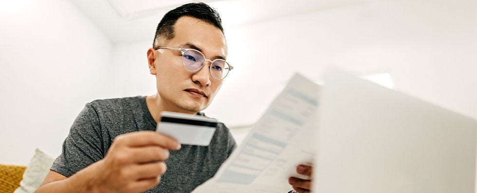 Man in glasses looking at credit card and paperwork in front of computer. 