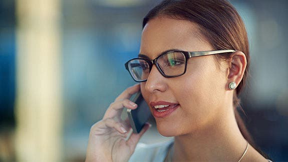 Photo of a woman speaking on a telephone in an office setting. She has glasses.