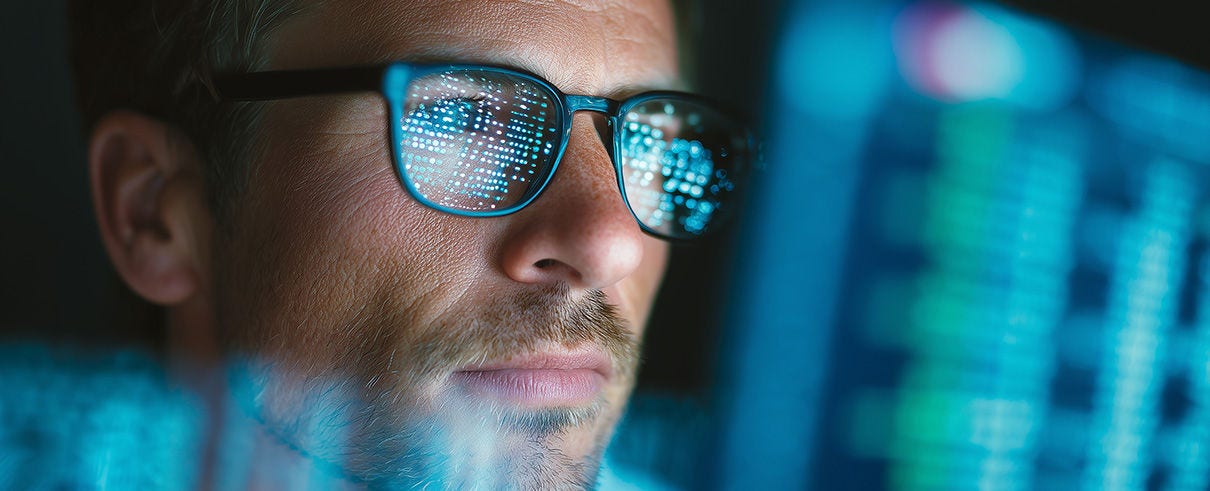 Close-up of a focused man wearing glasses reflecting digital code on multiple screens, working late on data analysis and software development in a dark modern office environment.