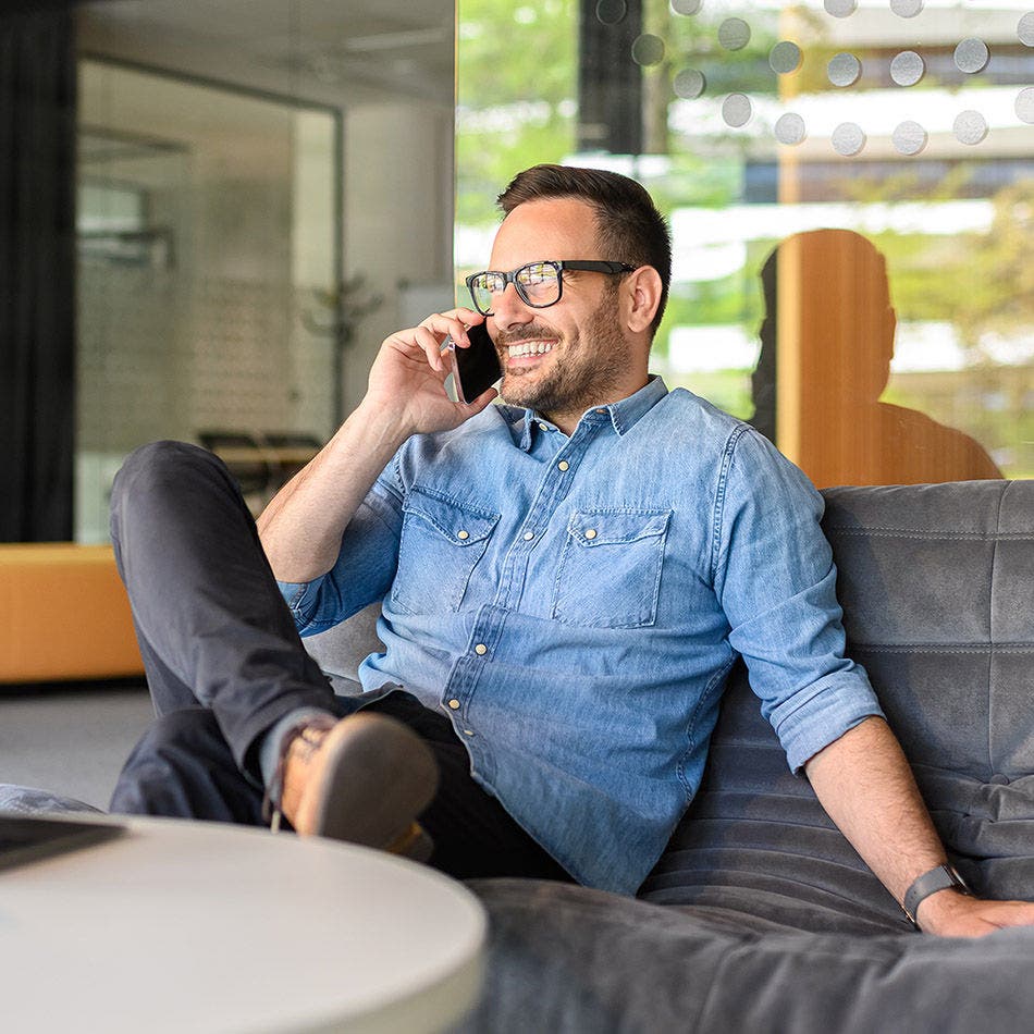 man wearing glasses smiling with laptop and holding phone