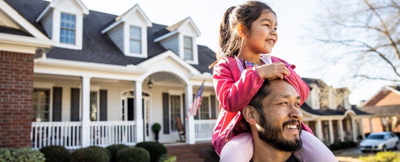 Father holding daughter on his shoulders in front of a home. 