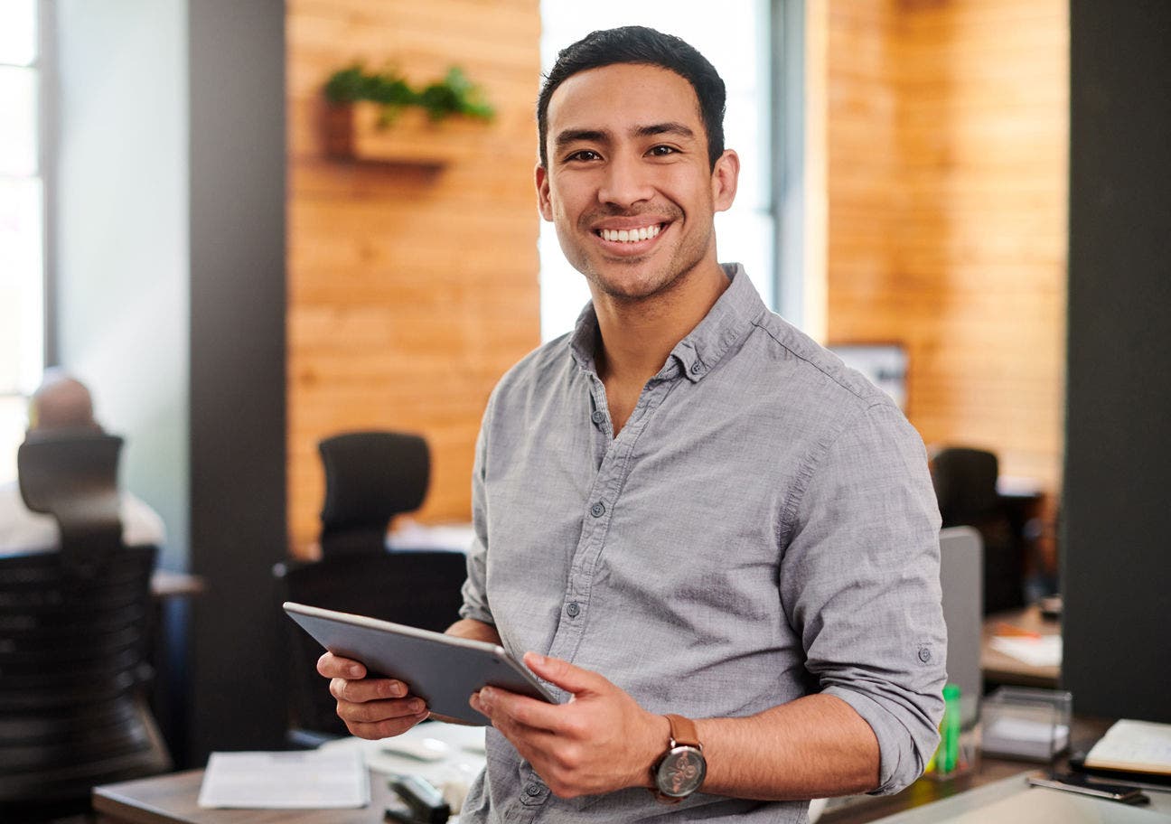 man in a casual office environment holding a tablet and smiling at the viewer