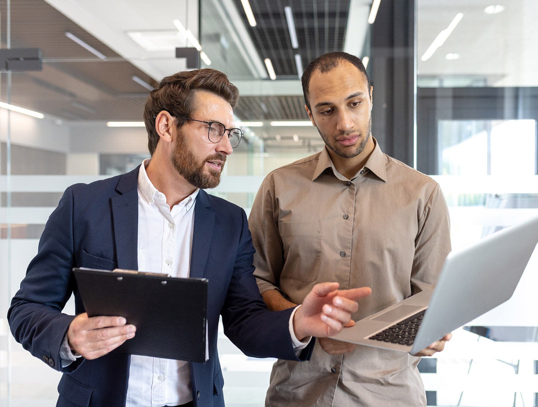 Two male colleagues review information on a laptop, collaborating inside an office setting for a business discussion.
