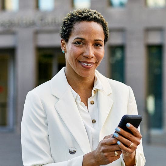 woman standing on the sidewalk using a smartphone and smiling at the viewer
