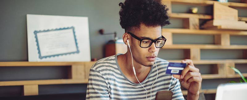 Man with headphones sitting and computer looking at the back of his credit card. 