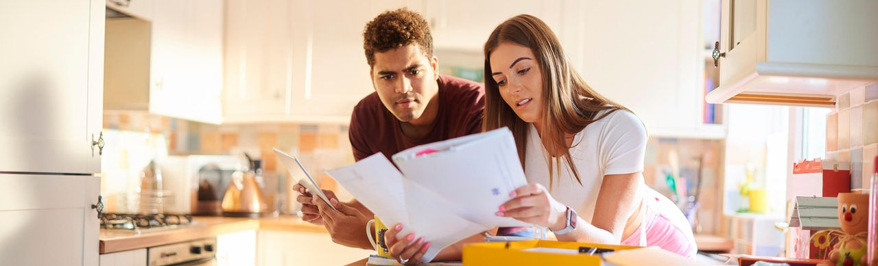 A lifestyle image showing two adults in a modern, well-lit kitchen reviewing paperwork that appears to include credit card statements and bills. One individual holds multiple envelopes and documents while the other looks at a tablet device. 