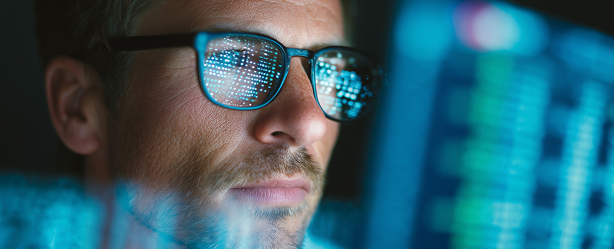 Close-up of a focused man wearing glasses reflecting digital code on multiple screens, working late on data analysis and software development in a dark modern office environment.