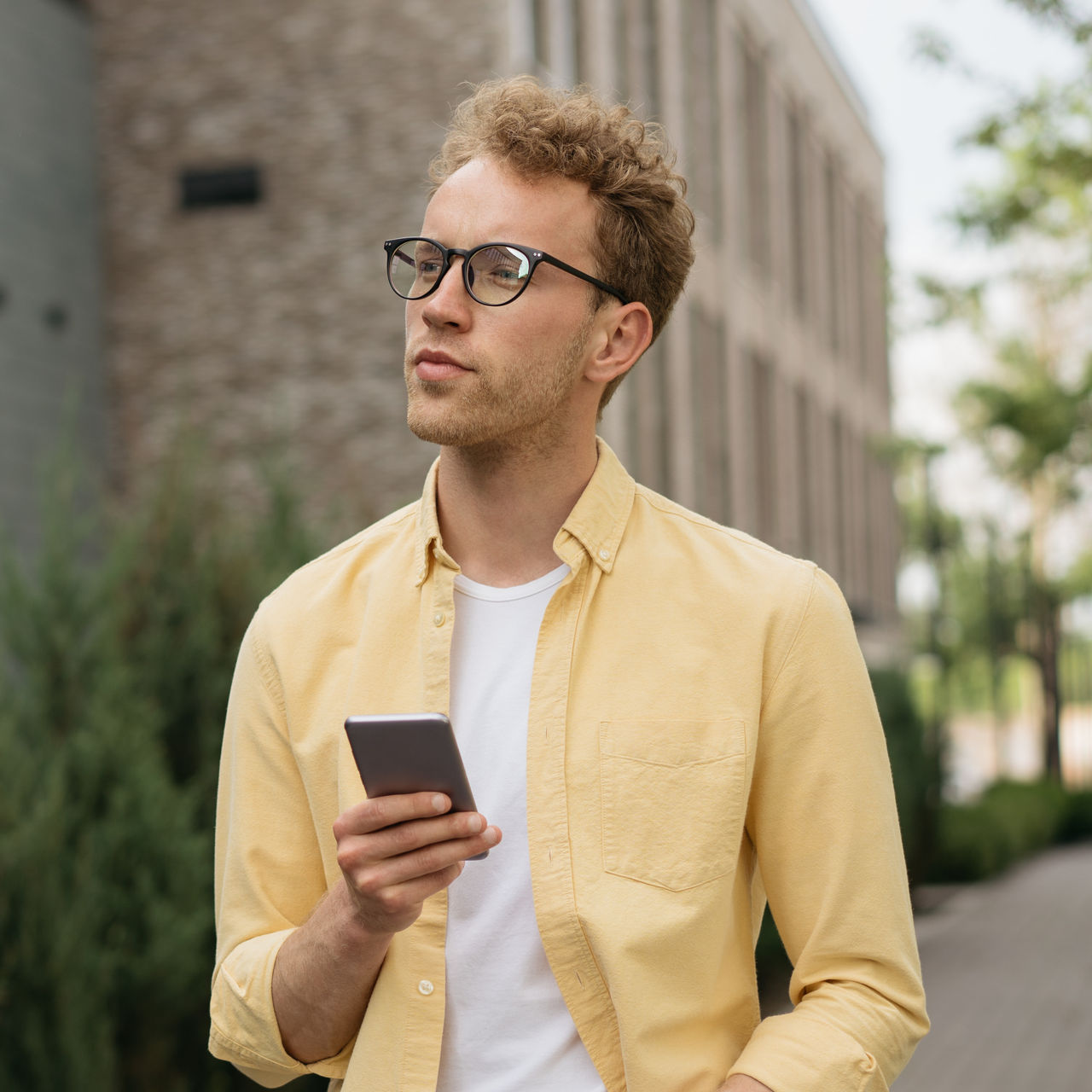Handsome pensive man wearing yellow casual shirt and stylish eyeglasses using mobile phone walking on the street, copy space