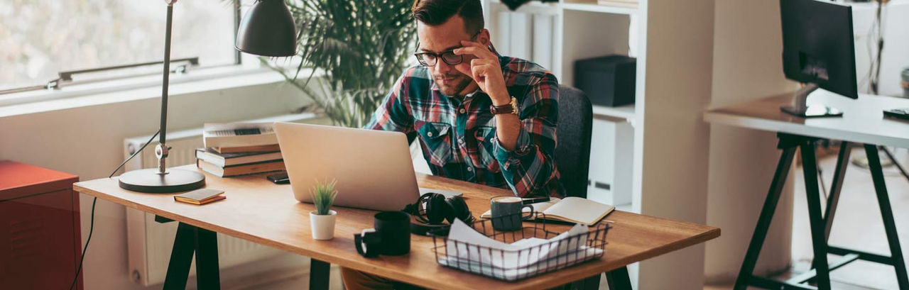 man wearing glasses sitting in home office looking at his laptop on desk. 