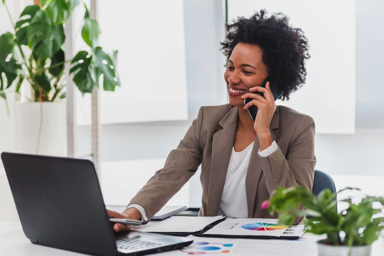 Happy smiling african-american business woman working on laptop at office, using smart phone. Businesswoman sitting at her working place